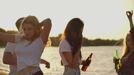 Three-female-students-with-perfect-bodies-are-dancing-in-short-t-shirts-with-beer-on-the-beach-party-at-sunset.-Their-long-hair-is-flying-on-the-wind.